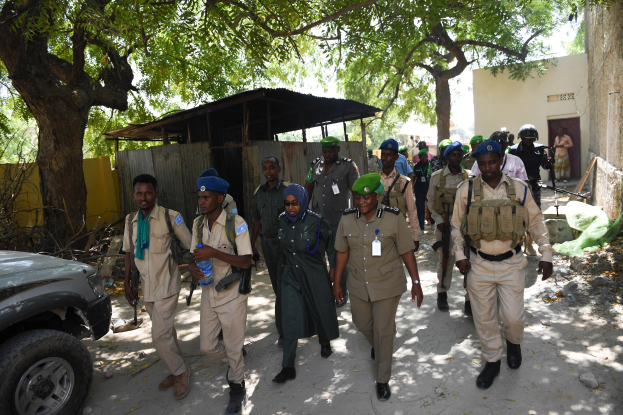 Eine Gruppe uniformierter Polizisten geht eine Straße entlang, einige tragen Waffen, mit Gebäuden, Bäumen und Schuppen im Hintergrund.