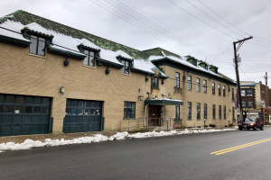Großes Backsteingebäude mit grüner Dachfläche an einer Straßenkreuzung, als ehemalige St. Louis-Feuerwache bezeichnet, umgeben von Fahrzeugen, Schnee, Strommasten, Bäumen und einem bewölkten Himmel.