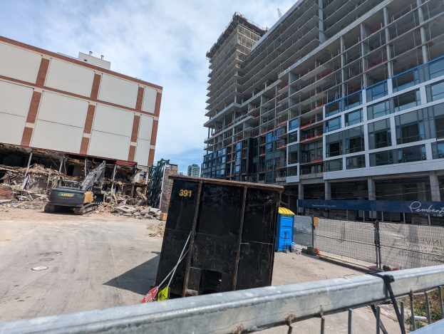 Ein großes Gebäude mit Schutt davor, umgeben von einem Metallzaun, einem Container, Pflanzen, Bäumen, Pfählen, einem Schild, Fahrzeugen, Steinen und einem bewölkten Himmel, was auf eine Baustelle für einen neuen Wohnkomplex hinweist.