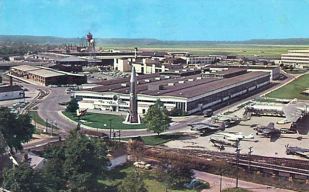 Altes Schwarz-Weiß-Foto eines Flughafens mit parkenden Flugzeugen, Gebäuden, Bäumen, Straßen, Fahrzeugen, Strommasten, Gras, Bahnschienen, Türmen, Hügeln und einem klaren blauen Himmel; Text erscheint unten.