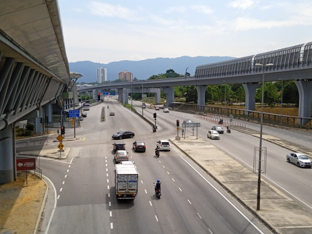 Eine vielbefahrene Autobahn mit mehreren Fahrzeugen, neben einer Brücke, mit Straßenlaternen, Verkehrsampeln, Verkehrsschildern, Bäumen, Gebäuden, Hügeln und einem bewölkten Himmel.