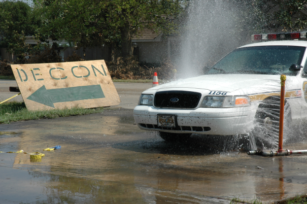 Ein Polizeiauto fährt durch eine Pfütze, mit einem Schild, einer Verkehrskegel und anderen Gegenständen auf dem Boden, mit Bäumen, Gebäuden und einem klaren blauen Himmel im Hintergrund.