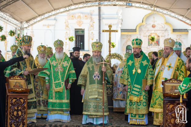 Gruppe von Priestern vor einer Kirche während einer religiösen Zeremonie, mit einem Buch und einem Mikrofon haltenden Person und einem Kreuzsymbol, Blumen und einem Gebäude im Hintergrund.
