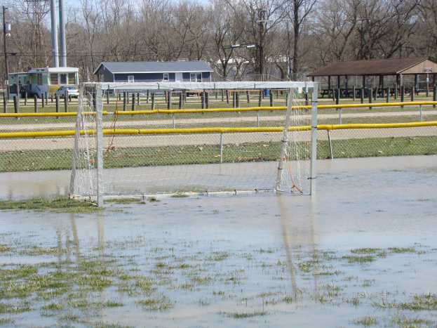 Ein Fußballtor steht in einem überfluteten Feld mit Gras und Wasser, umgeben von Hütten, Pfosten, Bäumen, Fahrzeugen und einem klaren blauen Himmel im Hintergrund.