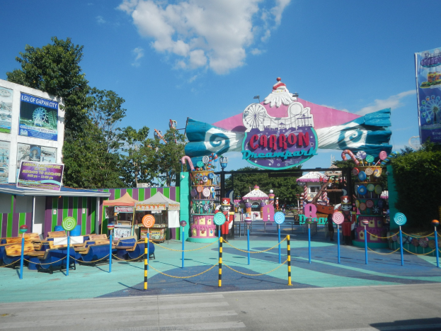 Karussell am Parkeingang mit Straße, Absperrung, Buden, Banner, Bäumen und bewölktem Himmel.