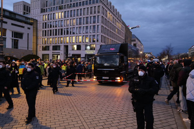 Eine Gruppe von Menschen steht vor einem Lkw auf einer Straße, umgeben von Gebäuden, Laternenmästen, Bäumen und einem bewölktem Himmel, einige tragen Mötzen und Masken, und ein Band mit einem Pfahl im Vordergrund.