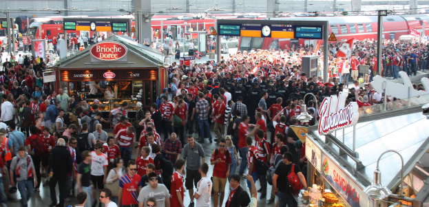 Eine große Menschenmenge bewegt sich durch einen belebten Bahnhof, im Hintergrund ist ein stehender Zug auf den Gleisen zu sehen.