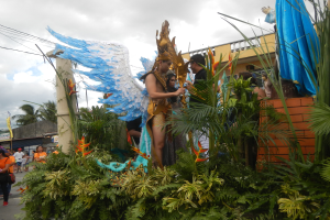 Eine Gruppe von Menschen geht eine Straße entlang neben einem Karnevalswagen, der mit Pflanzen und Blumen geschmückt ist, mit kostümierten Personen auf dem Wagen; Gebäude, Bäume und Strommasten mit Drähten sind im Hintergrund unter einem bewölkten Himmel sichtbar.