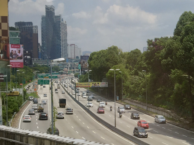 Eine belebte Autobahn mit hohen Gebäuden, Straßeninfrastruktur und Bäumen unter einem bewölkten Himmel.