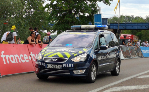 Polizeiauto fährt auf einer Straße neben einer Menschenmenge, mit einem Banner auf der linken Seite des Autos und Geländern mit Bannern dahinter, Bäumen und einer Brücke im Hintergrund.