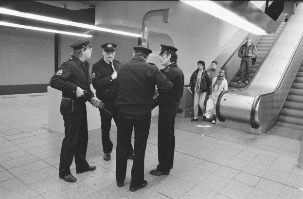 Schwarzes und weißes Bild von mehreren Polizeibeamten, die in einer U-Bahn-Station neben einer Rolltreppe mit einigen Menschen darauf stehen.