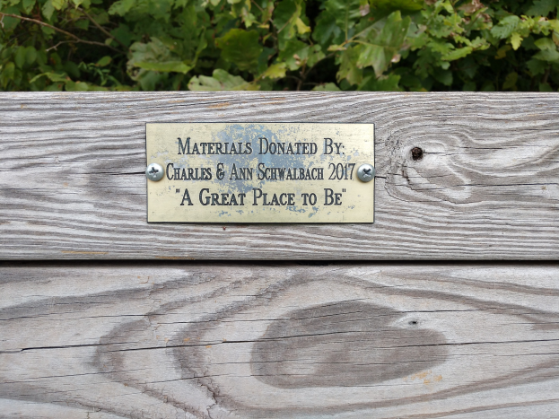 Holzbank mit einer Plakette, auf der "Materialien gespendet von Charles & Ann Schwabach 2017: Ein schöner Ort" steht, in einem Garten mit üppiger Vegetation.