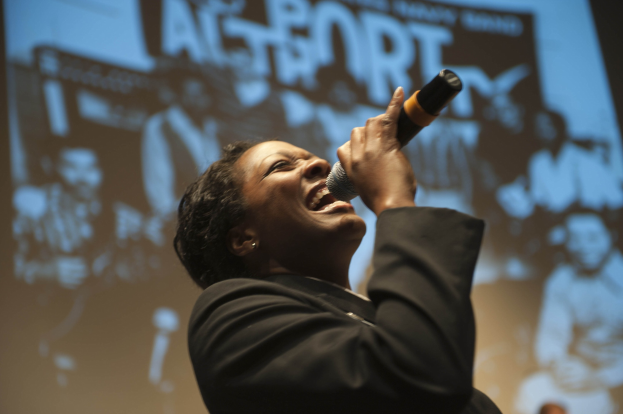 Eine Frau in einem schwarzen Kleid mit einem Dutt singt leidenschaftlich in ein Mikrofon auf der Bühne, ihr Gesicht von Scheinwerfern beleuchtet und ihre Augen vor Emotion geschlossen.