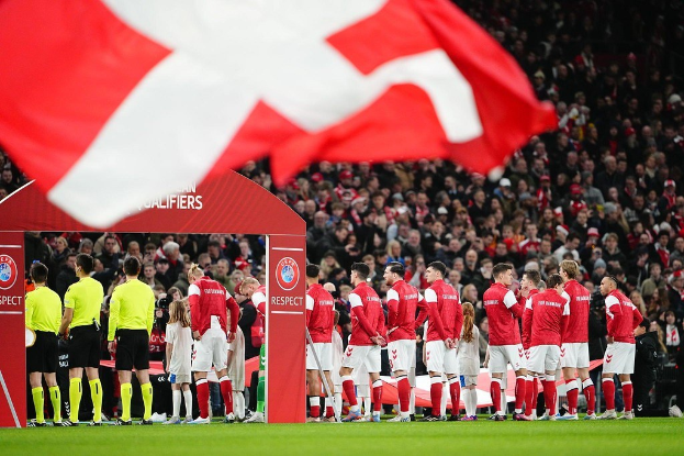 Eine Gruppe von Menschen auf einem Fußballfeld mit einer roten und weißen Flagge im Vordergrund, ein Bogen mit der Aufschrift "Bayern München vs. Bayern München Wetten & Vorschau" im Hintergrund und eine große Menschenmenge im Stadion.
