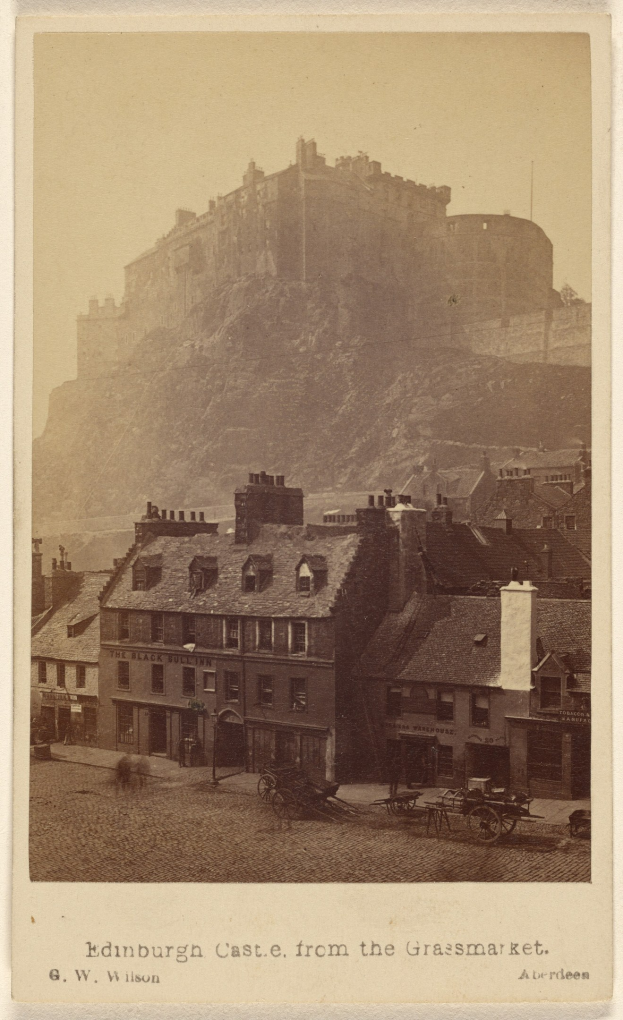 Alte Fotografie des Edinburgh Castle von der Grassmarket aus betrachtet, mit Gebäuden und Karren im Vordergrund und einem Hügel im Hintergrund, mit Text am unteren Rand.