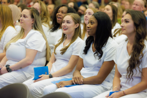 Eine Gruppe von Frauen in weißen Kleidern sitzt in einem Hörsaal, jede hält ein blaues Objekt, mit einem unscharfen Hintergrund, der die Frauen betont, die Teil eines Pflegeprogramms zu sein scheinen.