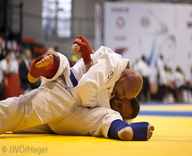 Zwei Judo-K├Ąmpfer im Zentrum einer Dojo mit Zuschauern und Ausrüstung im Hintergrund.