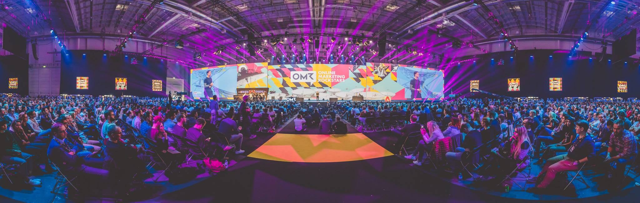 Eine große Zuschauermenge sitzt auf Stühlen einem hell erleuchteten Bühne mit mehreren Rednern und Bildschirmen gegenüber, was auf ein Online-Marketing-Event hindeutet.