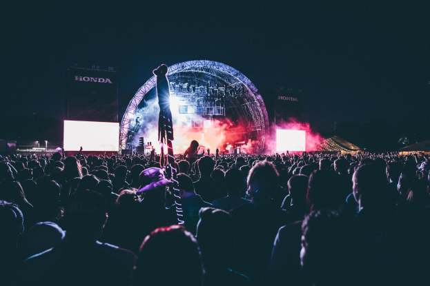 Eine große Menge bei einem Musikfestival mit einem Mann auf der Bühne umgeben von Musikern, beleuchtet von Lichtern, Bildschirmen und Bannern vor einem dunklen Hintergrund.