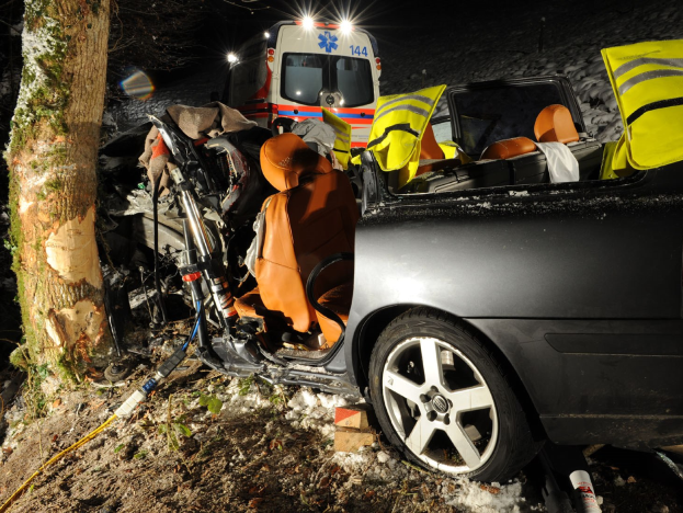 Ein geschädigtes Auto am Straßenrand mit einem Krankenwagen im Hintergrund, umgeben von Bäumen und Schnee.