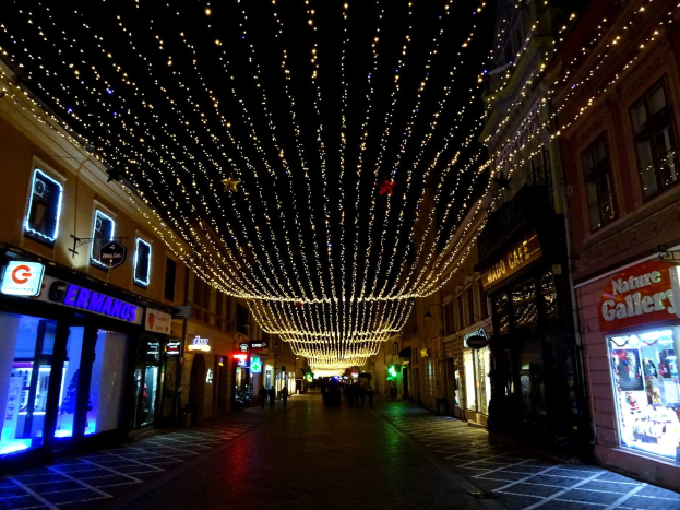 Eine nächtliche Stadtstraße mit Weihnachtsbeleuchtung, gesäumt von Gebäuden mit beleuchteten Fenstern und Schildern, mit einigen Passanten auf dem Weg.