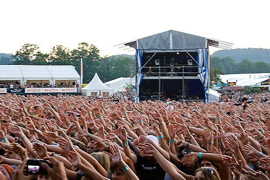 Eine große Menschenmenge auf einem Musikfestival mit erhobenen Händen und Handys, eine Bühne im Hintergrund, weiße Zelte, Bäume und einen klaren blauen Himmel.