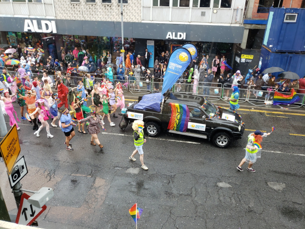 Eine Gruppe von Menschen in bunter Kleidung, die eine Straße in einer Parade entlanggehen, mit einem Auto in der Mitte, einige halten Schirme, und Gebäude im Hintergrund.