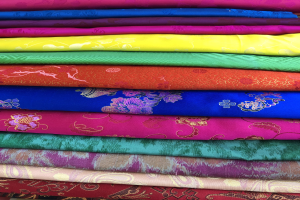 Ein farbenfroher Anblick von ordentlich angeordneten Stoffen auf einem Markt, mit leuchtenden Blüten, Grünen, Orangen und Gelb.