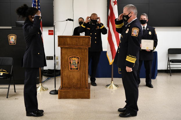 Gruppe uniformierter Personen in Masken auf einem Podium mit einem Mikrofon, umgeben von Fahnen und Möbeln, während einer Vereidigung eines neuen Feuerwehrchefs.