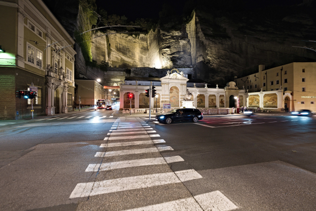 Eine nächtliche Stadtstraße mit fahrenden Autos, Straßenlaternen, Verkehrsampeln, beleuchteten Fenstern in Gebäuden, Laternenpfählen, einem Hügel im Hintergrund und einer Zebrastreifenfurt am unteren Bildrand.