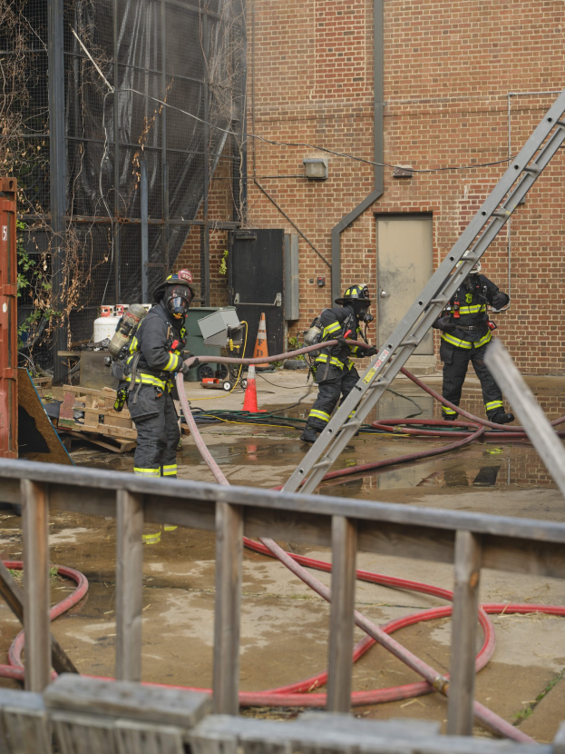 Firefighters in helmets work to extinguish a building fire, surrounded by equipment and a metal fence, with a tree and sky in the background.