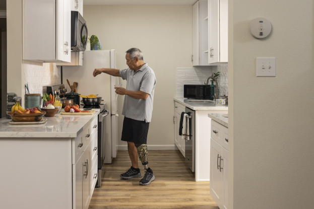Ein Mann steht in einer Küche mit weißen Schränken, hält ein Glas in der Hand und ist von Obst, Gemüse, Schüsseln und Küchenutensilien auf einer Plattform zu seiner Linken umgeben, mit Geräten und Schränken im Hintergrund.