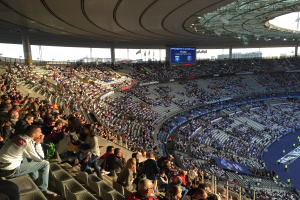 Große Menschenmenge in einem Stadion bei einem Fußballspiel mit einer Bühne, Fahnen, Stangen, einem Bildschirm und der Allianz Arena in München, Deutschland im Hintergrund.