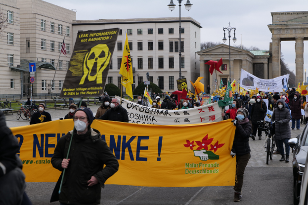 Große Gruppe von Menschen bei einer Straßendemonstration gegen Atomkraft in Deutschland, mit Schildern und Fahnen, sowie Fahrzeugen, Gebäuden, Bäumen und einem Tor im Hintergrund.