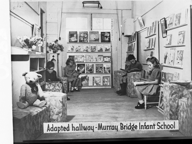 Gruppe von Kindern auf Stühlen in einem bibliotheksähnlichen Raum mit Bücherregalen, einem Teddybär links, einer Blumenvase auf dem Tisch, gerahmten Gegenständen an der Wand und Text unten mit "Angepasster Flur Murray Bridge Kindergarten"