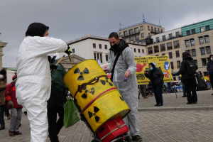 Eine Gruppe von Menschen in weißen Anzügen und Masken steht um einen gelben Bottich mit einem Wagen davor, mit Schildern in der Hand, vor Gebäuden mit Fenstern und Fahnen, unter einem bewölkten Himmel.