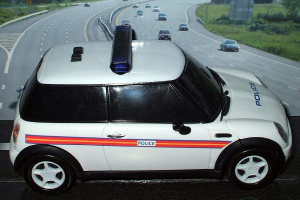 Weißes Polizeiauto vor einer Reihe von fahrenden Fahrzeugen auf einer von Gras und Bäumen gesäumten Straße ausserhalb der Stadt.