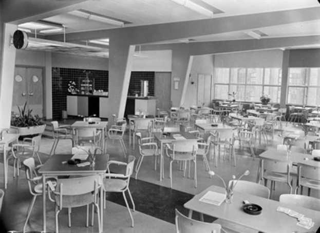 Schwarz-weißes Foto einer Cafeteria mit Tischen und Stühlen, Tellern, Besteck, Gläsern und Blumenvasen auf den Tischen und einer Wand mit Türen, Fenstern und Deckenleuchten im Hintergrund.