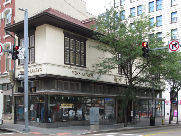 Eine belebte Stadtstraße mit einem geparkten Bus, Gebäuden, Bäumen, Laternen, Verkehrsampeln, Schildern und einem Kaufhaus mit Schaufensterpuppen in Kleidern, mit dem Himmel im Hintergrund.