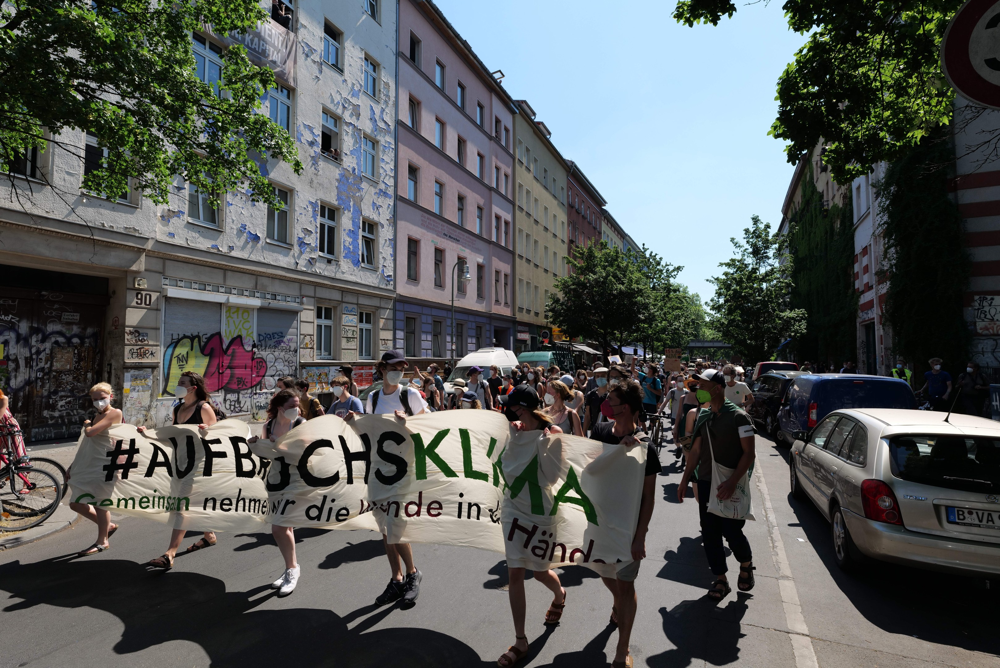 Eine Gruppe von Menschen marschiert bei einer Demonstration, hält ein Banner in der Hand, mit einem Radfahrer auf der linken Seite und parkenden Autos auf der rechten Seite, vor einer Kulisse aus Gebäuden, Bäumen und einem Schild in Berlin.