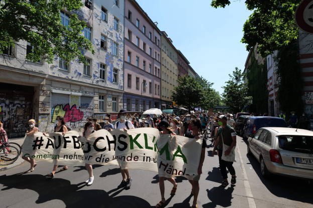 Eine Gruppe von Menschen marschiert bei einer Demonstration, hält ein Banner in der Hand, mit einem Radfahrer auf der linken Seite und parkenden Autos auf der rechten Seite, vor einer Kulisse aus Gebäuden, Bäumen und einem Schild in Berlin.