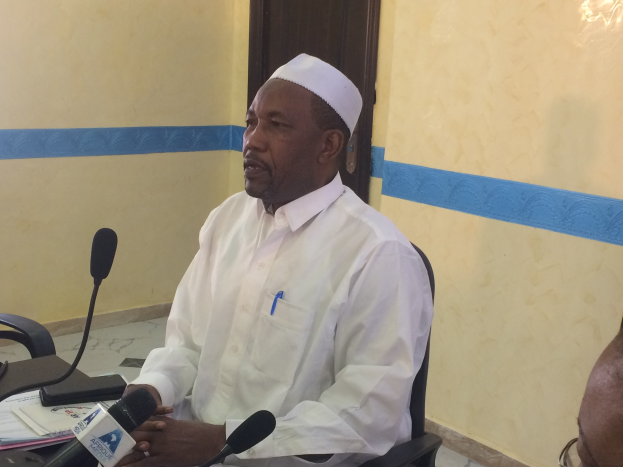 Ein Mann in weißem Anzug und Hut sitzt an einem Tisch mit Mikrofonen, spricht, während Papiere und Gegenstände auf dem Tisch sichtbar sind, mit einem anderen Mann mit Brille zu seiner Rechten, in einem Raum mit einer Wand und einer Tür im Hintergrund.