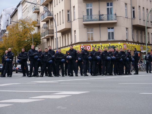 Eine Gruppe von Polizisten in schwarzen Uniformen mit blauen Helmen steht in der Mitte einer Straße, die von Laternenpfählen, Bäumen und Gebäuden mit Glasfenstern und Balkonen gesäumt ist, mit weiteren Bäumen und einem klaren blauen Himmel im Hintergrund.