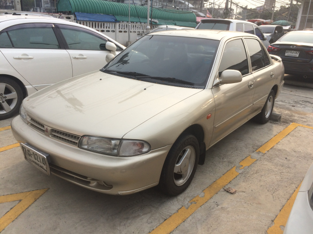 Ein goldfarbener Wagen auf einem Parkplatz mit anderen Autos, Gebäuden, Pfählen, Drähten, Bäumen und einem klaren blauen Himmel.