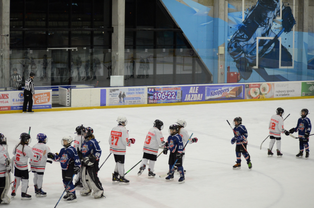 Gruppe junger Menschen beim Eisschnellaufen auf einer Indoor-Eisbahn, die Helme tragen und Stöcke halten, mit einer Person links und Bannern, Säulen und Glaswänden im Hintergrund.