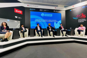 Eine Gruppe von Menschen sitzt auf einer Bühne beim Östlichen Wirtschaftsforum in Moskau, mit Flaschen und anderen Gegenständen auf den Tischen, Bannern mit Text und Logos im Hintergrund und Deckenbeleuchtung.