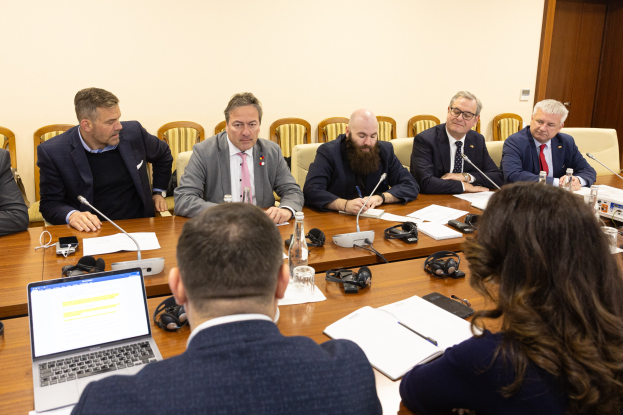 Eine Gruppe von Menschen, wahrscheinlich Mitglieder des Europäischen Parlaments, sitzt um einen Tisch in einem Konferenzraum, umgeben von Laptops, Büchern, Papieren, Flaschen, Gläsern, Headsets, Mikrofonen mit Ständern und Mobiltelefonen, mit einer Wand und einer Tür im Hintergrund.