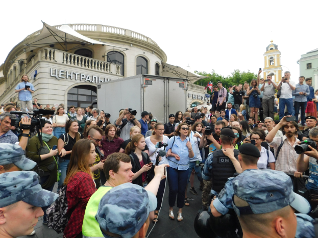 Eine große Gruppe von Menschen steht vor einem Gebäude, einige halten Kameras und Telefone, mit einem geparkten Fahrzeug, Bäumen und einem klaren blauen Himmel im Hintergrund, wahrscheinlich protestieren sie gegen das Verbot von Drohnen durch die russische Regierung.
