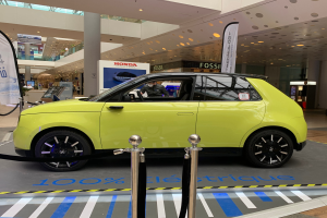 Ein gelbes wasserstoffbetriebenes Elektroauto vor einem Gebäude geparkt, umgeben von Absperrpollern mit Seilen und Bannern, mit einer Person darin, einer Säule, einer Gruppe von Menschen, Schildern, einer Topfpflanze, Deckenleuchten und einer Decke mit Deckenventilatoren.