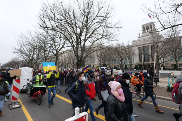 Eine große Gruppe von Menschen nimmt an einer Protestdemo in Washington, D.C. am 21. Januar 2020 teil, geht eine Straße entlang, hält Schilder und Banner hoch und fährt Fahrräder, während im Hintergrund Bäume und Schilder zu sehen sind und der Himmel klar blau ist.